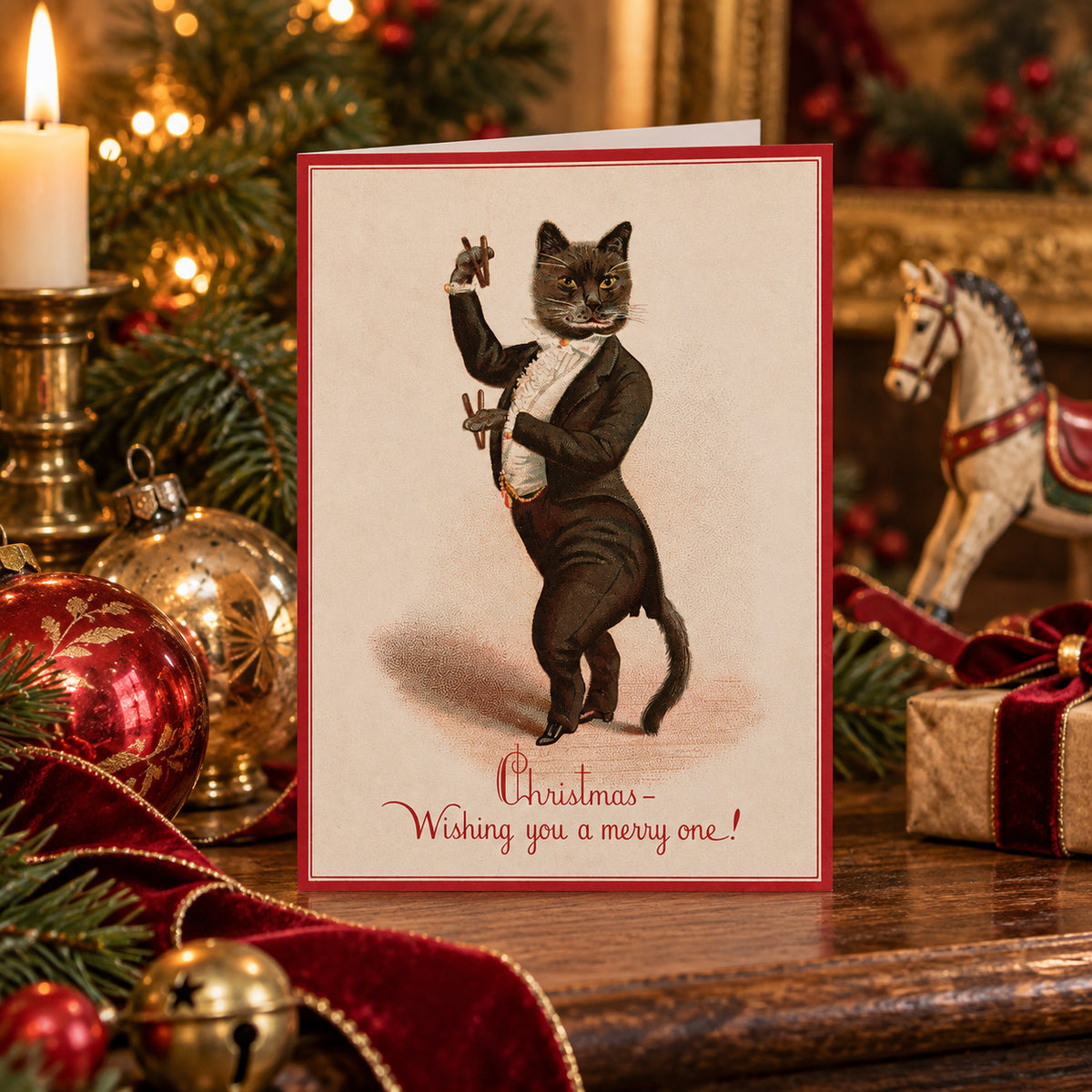 A Christmas greeting card featuring a vintage-style illustration of an anthropomorphic black cat in a tuxedo holding castanets, with the text 'Christmas - Wishing you a merry one!' at the bottom. The "weird Victorian" greeting card is displayed on a wooden surface surrounded by holiday decorations, including a lit candle, red ornaments, green pine branches, and a horse figurine.
