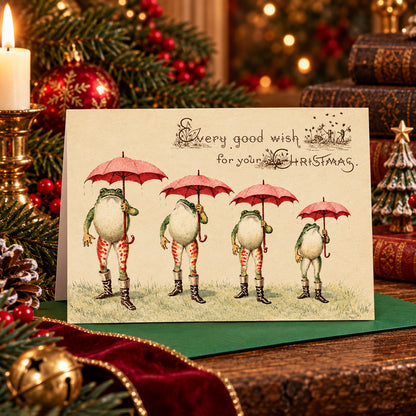 A  "weird Victorian" Christmas greeting card featuring four frogs in boots holding red umbrellas, with the text 'Every good wish for your CHRISTMAS.' The card is displayed in a festive holiday setting with pine branches, red ornaments, a candle, and books in the background.