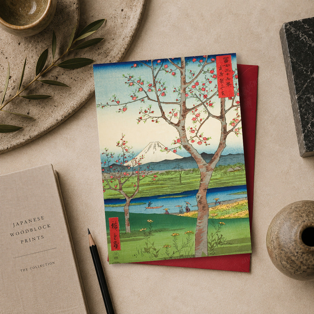A Japanese woodblock-style art Greeting card featuring cherry blossoms, a river, and Mount Fuji, styled on a beige surface with a red envelope, a pencil, a book, and decorative objects. “Koshigaya, Musashi Province” (武蔵越かや在 / Musashi Koshigaya-zai), 1858
by Utagawa Hiroshige (1797–1858), from the series Thirty-six Views of Mount Fuji.