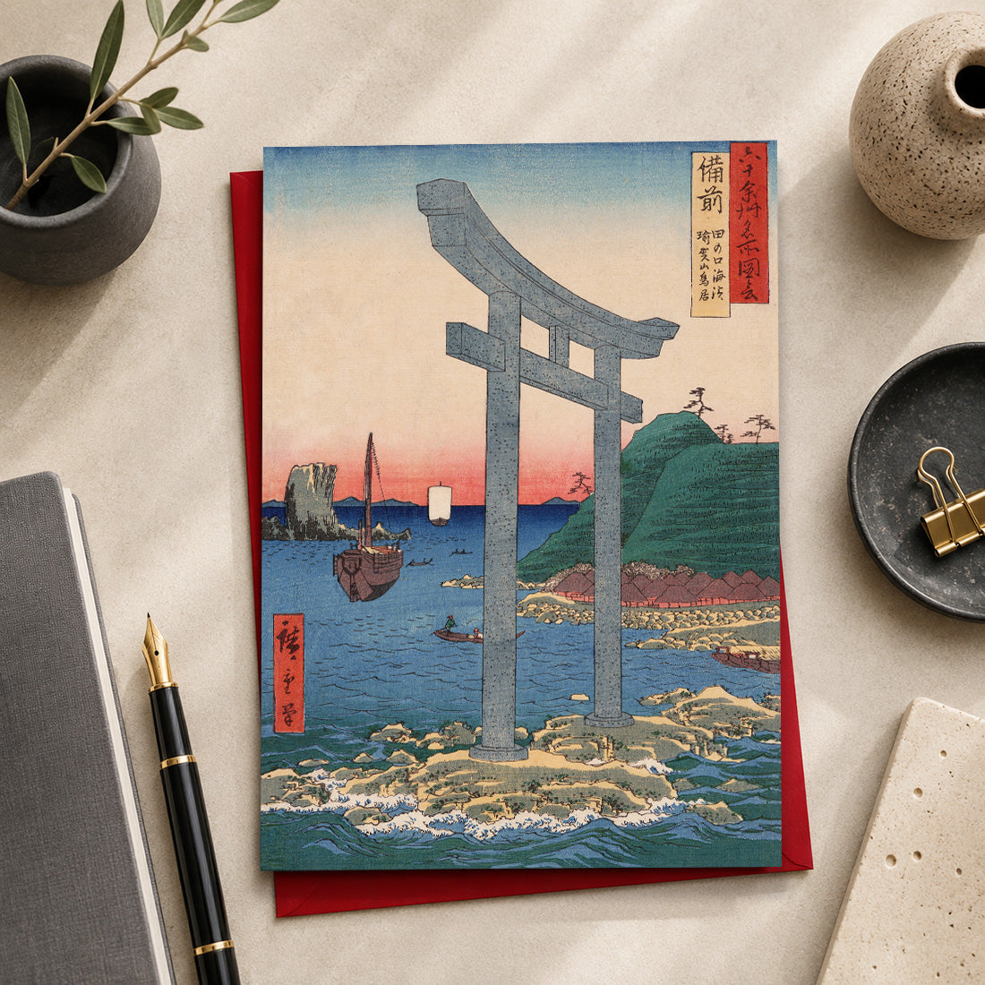 A Japanese-art greeting card featuring a large torii gate over blue water, placed on top of a red envelope or card. The print is photographed in a styled desk setting with a notebook, pen, ceramic dish, and small plant. The art featured is “Torii Gate to Mount Yuga Shrine at Tanokuchi Coast, Bizen”
(備前 田の口海浜 瑜賀山鳥居 / Bizen Tanokuchi Kaihin Yuga-san Torii), 1853
by Utagawa Hiroshige (1797–1858), from the series Famous Places of the Sixty-odd Provinces.