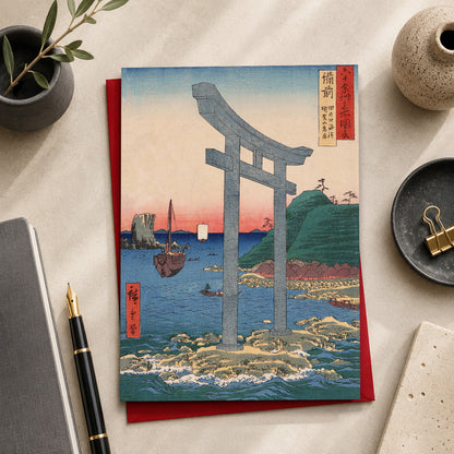 A Japanese-art greeting card featuring a large torii gate over blue water, placed on top of a red envelope or card. The print is photographed in a styled desk setting with a notebook, pen, ceramic dish, and small plant. The art featured is “Torii Gate to Mount Yuga Shrine at Tanokuchi Coast, Bizen”
(備前 田の口海浜 瑜賀山鳥居 / Bizen Tanokuchi Kaihin Yuga-san Torii), 1853
by Utagawa Hiroshige (1797–1858), from the series Famous Places of the Sixty-odd Provinces.
