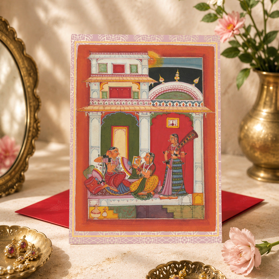 A large Indian art greeting card featuring Vilaval Ragini, ca. 1680, from a Ragamala series — a painted Garland of Musical Modes.
In this intimate morning scene, an elegantly dressed woman prepares to meet her lover, adjusting her earrings as a companion holds up a mirror beside her. Rich reds, oranges and greens, patterned surfaces and jewel-like architectural details turn a quiet moment of anticipation into something wonderfully sumptuous.